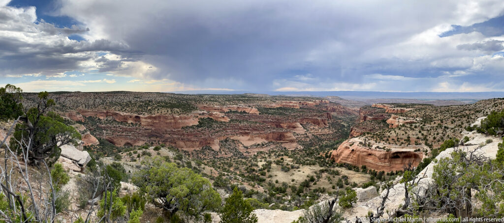 A broad red sandstone canyon spreads below a rocky overlook as dark clouds release rain in the distance.