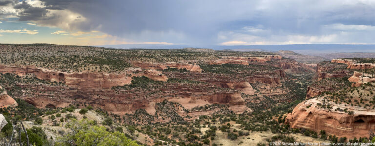 A wide canyon of red cliffs and pale benches stretches to the horizon beneath clouds and falling rain.
