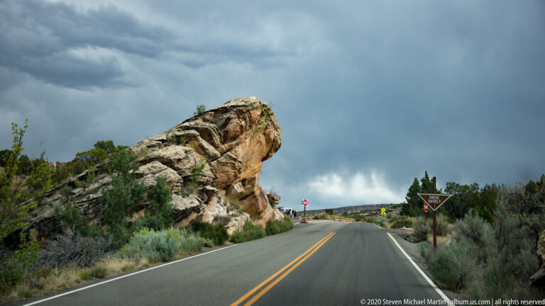 A highway approaches a signed junction beside a massive tilted boulder and low shrubs beneath storm clouds.