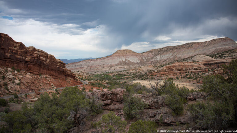 Red canyon ledges and scrub lead toward a pale layered mesa beneath heavy clouds and a distant bright horizon.
