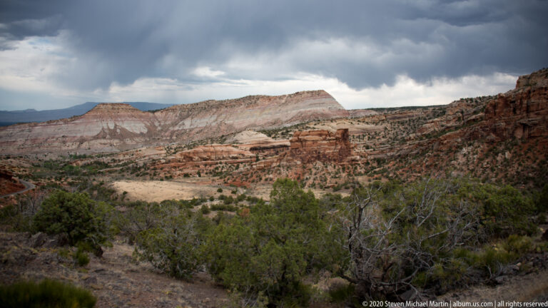 A pale flat-topped mesa rises behind red canyon cliffs and scrub under a thick cloudy sky.