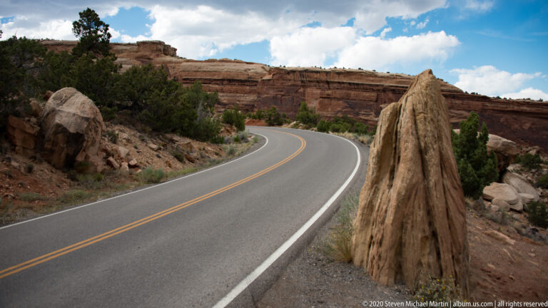 A curving two-lane highway passes a tall narrow sandstone fin, with layered cliffs and shrubs nearby.