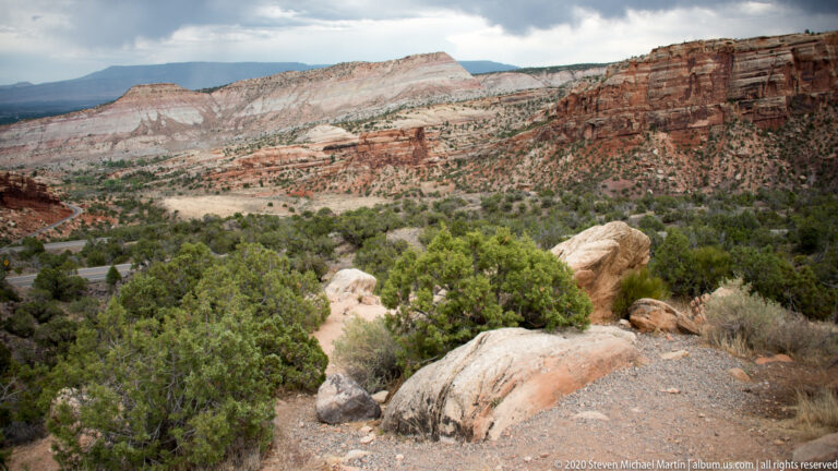A rocky footpath descends through shrubs and pale boulders toward a road below layered canyon cliffs.