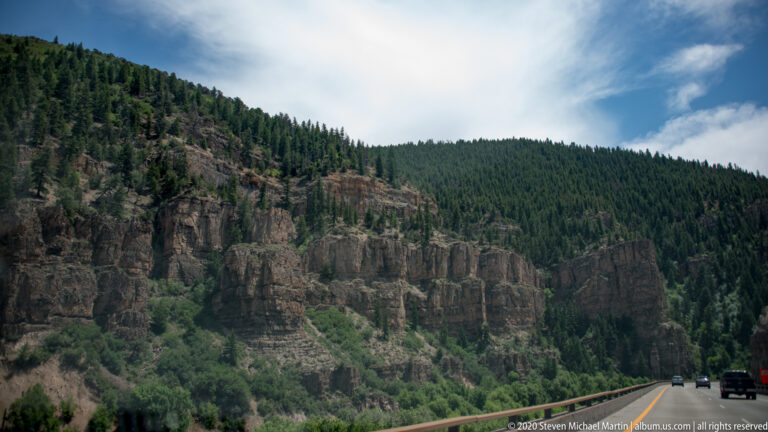 A highway with several vehicles runs below layered canyon cliffs covered in evergreen trees and shrubs.