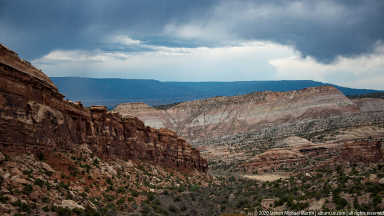 A deep canyon valley opens between a dark red wall and a pale layered mesa beneath broken clouds.