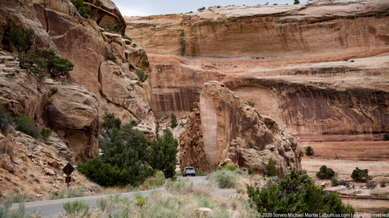A small vehicle rounds a highway beneath tall sandstone walls with a broad recess in the cliff face.