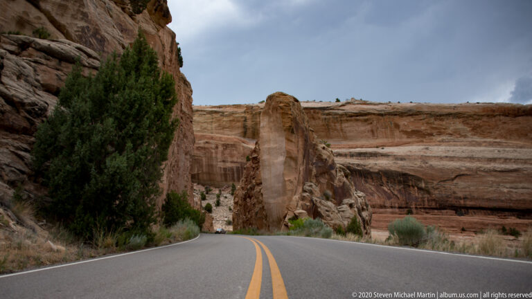 A two-lane highway leads between tall sandstone fins, with double yellow lines centered in the foreground.