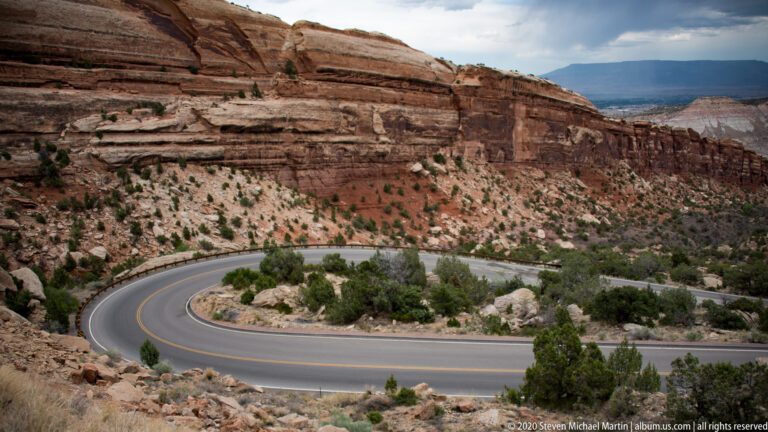 A highway hairpin curves beneath layered sandstone cliffs, edged by a guardrail and scattered shrubs.