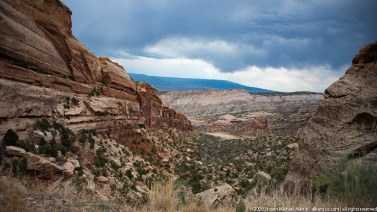 Rock walls frame a distant pale mesa and broad canyon valley beneath a clouded sky.