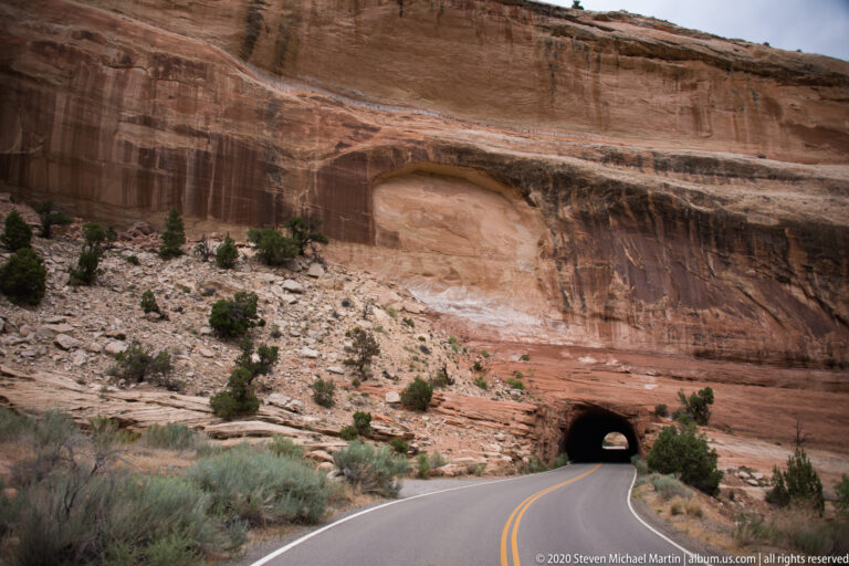 A curving highway approaches a dark tunnel portal beneath a broad sandstone wall with a shallow recess.