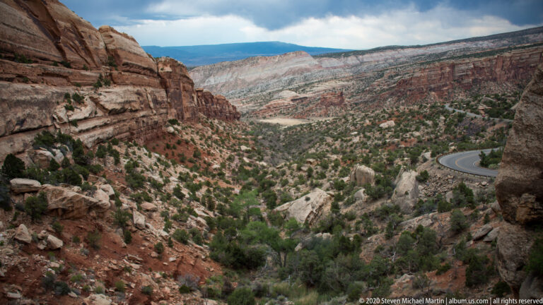 A high overlook looks across a layered canyon where a small road curves along the right slope.