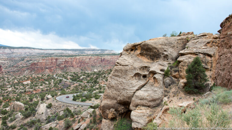 A pale pitted sandstone outcrop overlooks a curving road and distant red canyon cliffs.