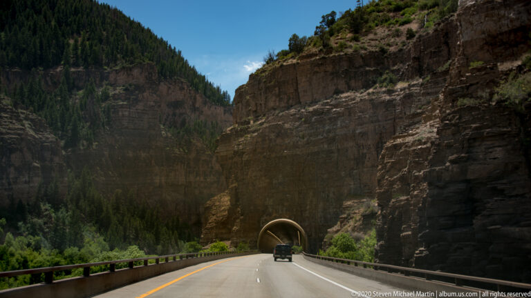 A vehicle approaches a dark tunnel cut through high canyon walls, following a paved road with a yellow center line.