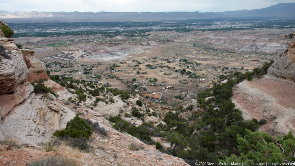 A rocky overlook looks across a broad valley with fields, roads, scattered buildings, and distant mesas.