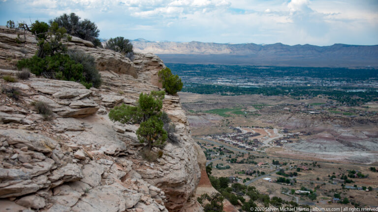 A pale sandstone ledge with junipers overlooks a broad valley, roads, and distant mesas.
