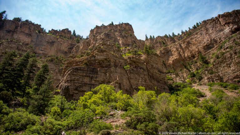 Sunlit sandstone cliffs rise above green shrubs and trees, with thin utility lines crossing the canyon view.