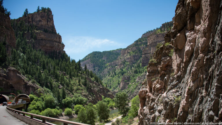 A curving highway passes a tunnel beside a river, with a guardrail and steep canyon walls framing the route.