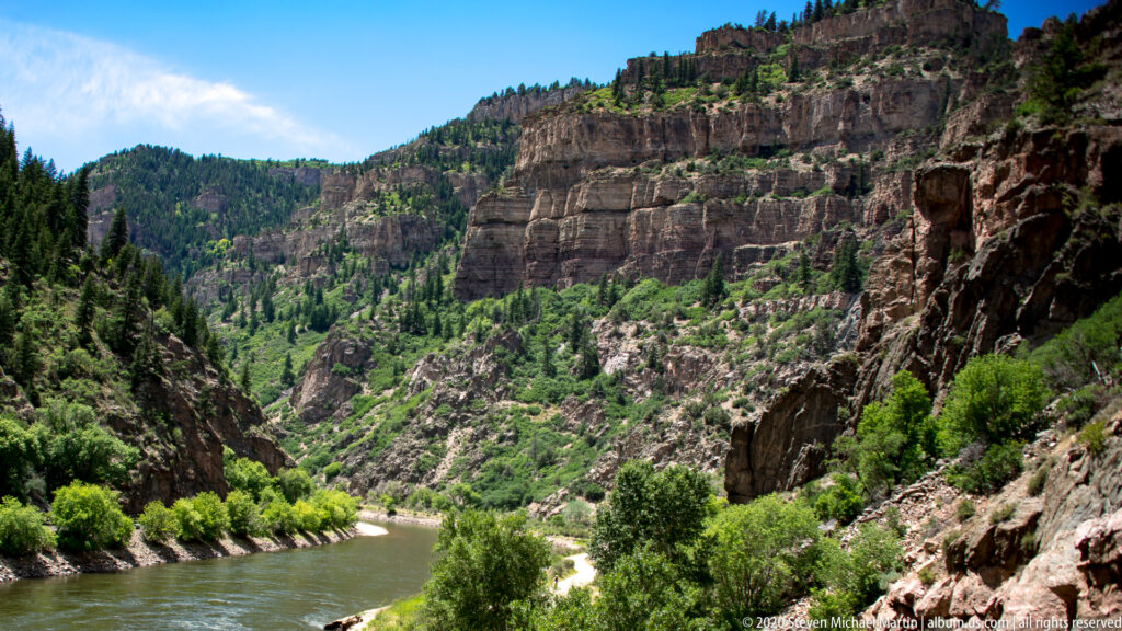 A river bends below layered canyon cliffs, with shrubs along the bank and rocky slopes rising on both sides.