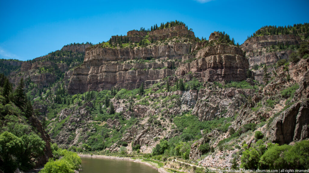 A green river winds through a canyon beneath a broad layered cliff, framed by rocky slopes and trees.
