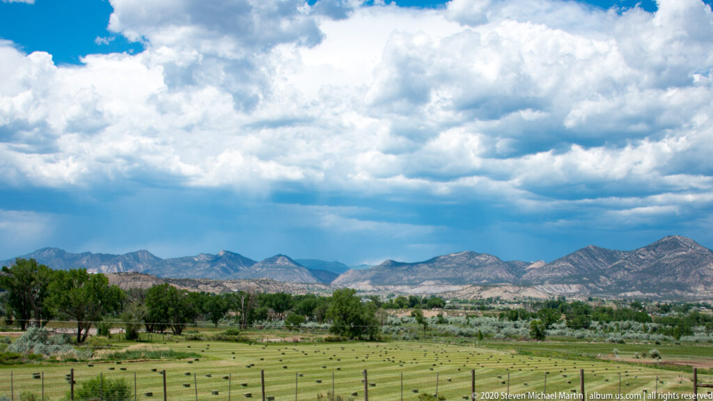 Rows of hay bales cover a fenced green field, with trees and a mountain range beneath large clouds.
