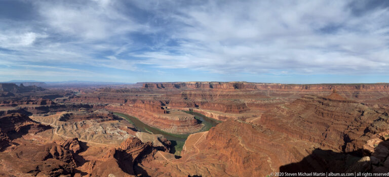 A broad Colorado River bend winds between red layered canyon walls and distant mesas beneath a cloud-streaked sky.