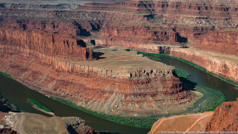 A green river curves around a broad sandstone peninsula beneath steep, terraced red canyon walls.