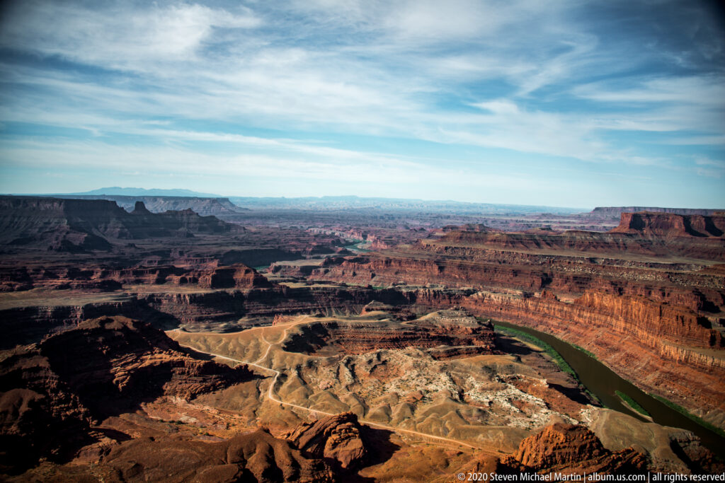 A wide plateau panorama shows layered mesas, branching ravines, and a distant green curve of the Colorado River.