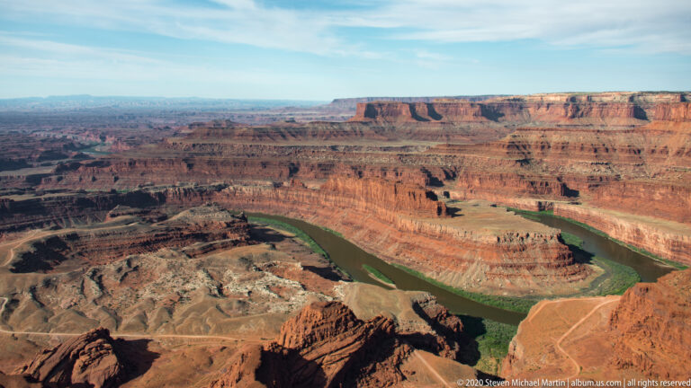 The Colorado River sweeps through a broad red-rock basin beneath a long mesa rim and pale terraces.