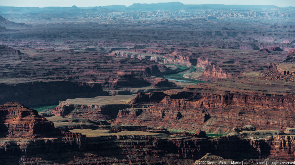 Shadowed canyon ledges lead toward distant mesas, with the Colorado River visible as a thin green line.
