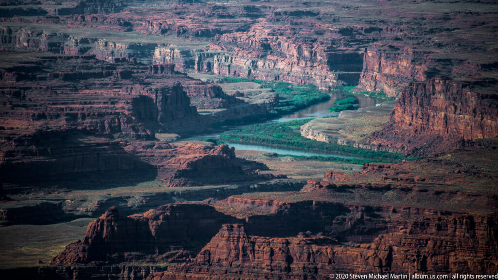 A green river threads between dark canyon walls and distant sunlit mesas beneath a hazy sky.