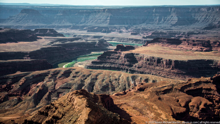 A winding river lies beyond pale eroded terraces and red ledges beneath a long distant cliff band.