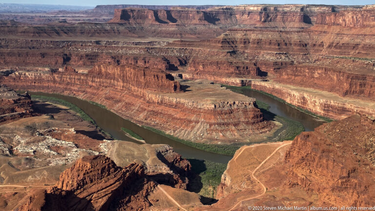 A green river curves around a pale sediment bench below steep, horizontally layered red canyon walls.