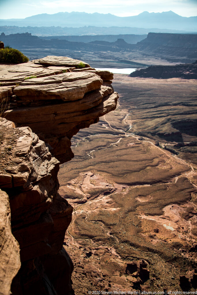 Thin sandstone shelves project over a broad arid basin with pale channels and distant hills.