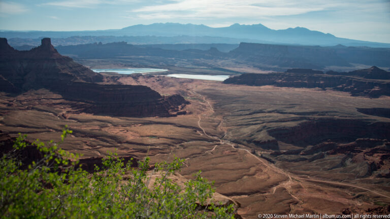 Pale drainage channels cross a broad canyon basin toward distant water, framed by dark mesas and hazy ridges.