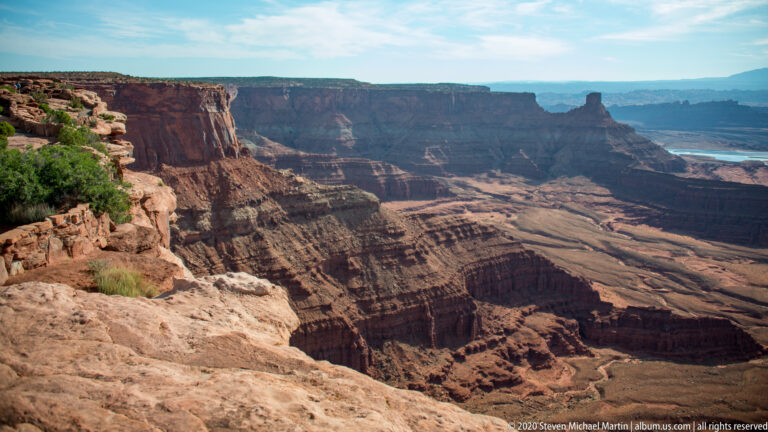 A rocky rim with sparse grasses overlooks deep red cliffs and a distant bend in the Colorado River.