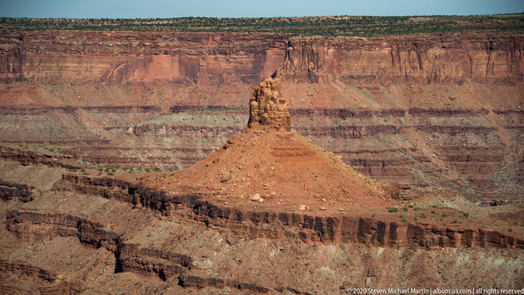 A narrow sandstone pinnacle rises from a red slope before a broad, layered canyon wall.