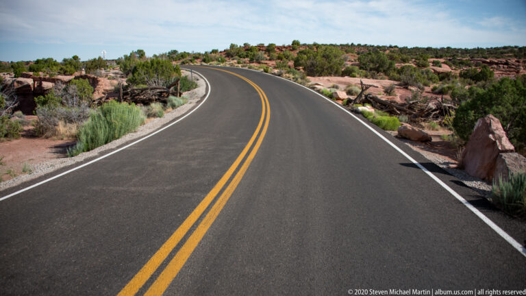 A paved two-lane road with double yellow lines curves between red sandstone shelves and desert shrubs.