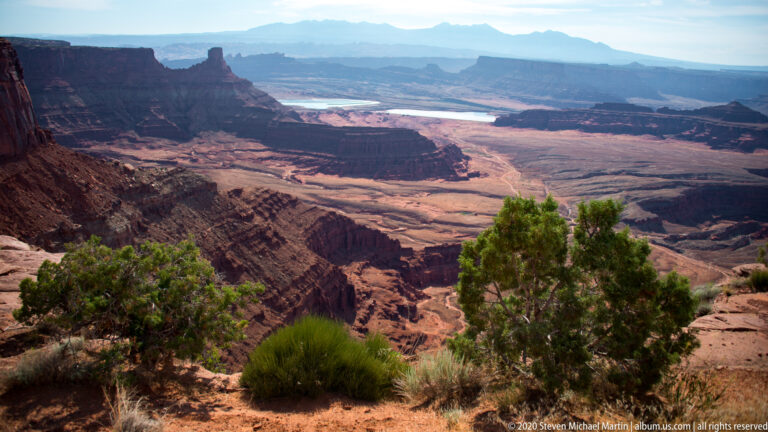 Junipers and low shrubs line a sandstone rim above a canyon, distant mesas, and a bright river bend.