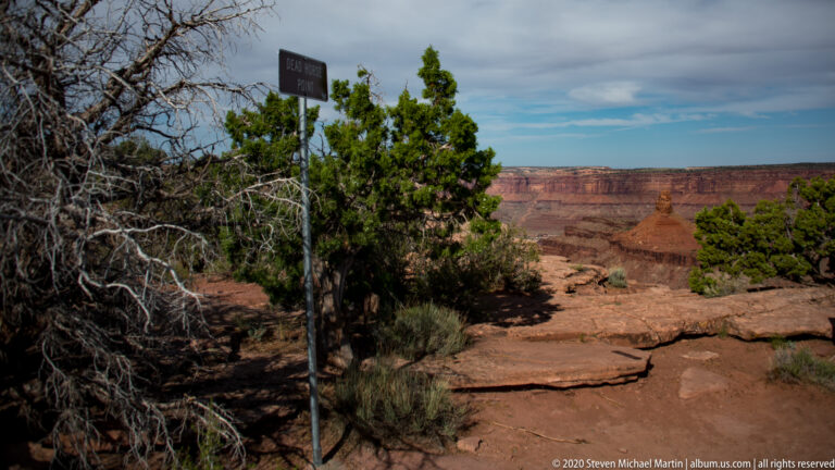 A dark signpost and junipers stand above red canyon walls and layered sandstone.