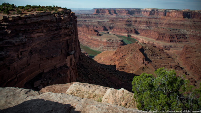 A green river winds through a terraced red-rock canyon beyond a pale ledge and shadowed cliff.