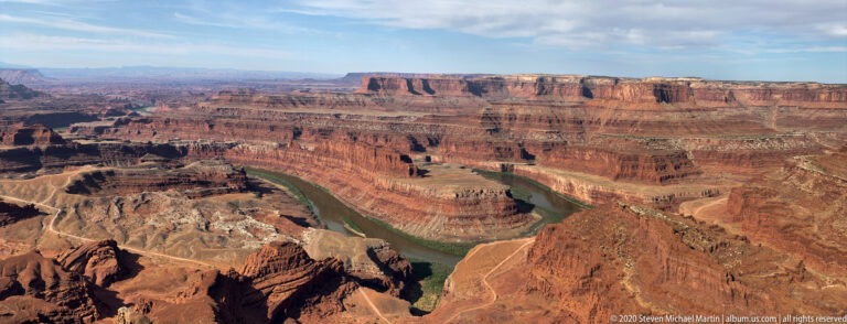 The Colorado River arcs through red cliffs and pale canyon benches beneath a distant flat-topped mesa.