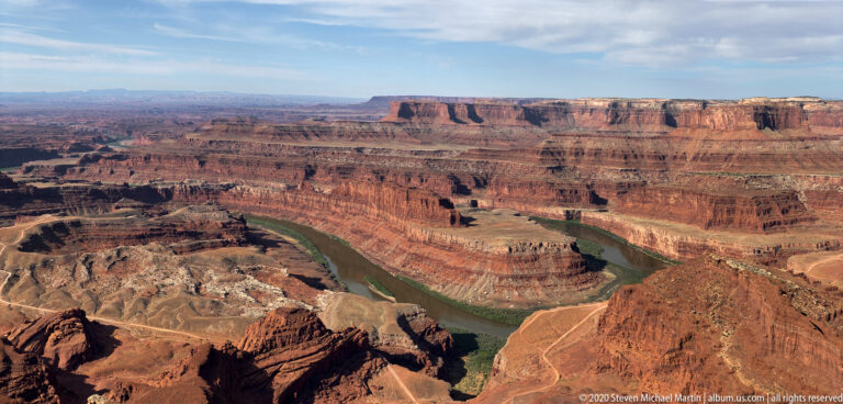 A green river loops around a rocky peninsula below a long red canyon rim and distant mesas.