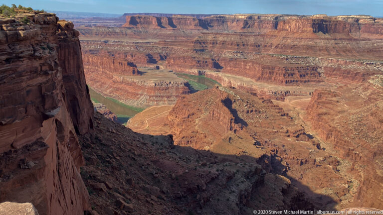A dark cliff frames the left side of a sunlit canyon where the Colorado River bends around red rock.