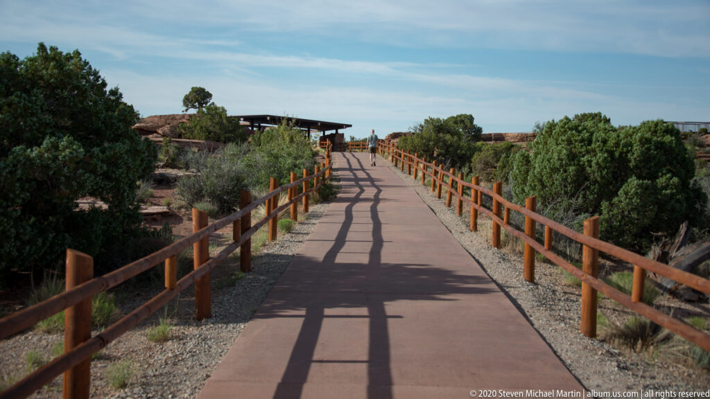A paved walkway with wooden railings leads through desert shrubs toward a shaded visitor structure.