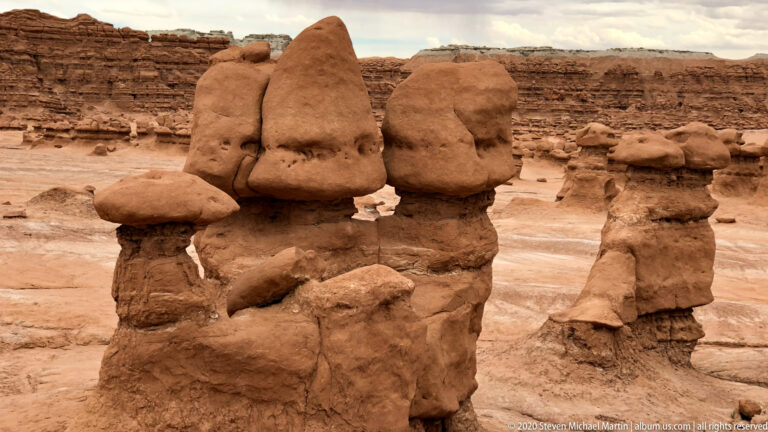 Rounded sandstone caps sit on narrow pedestals in a basin below a banded cliff.