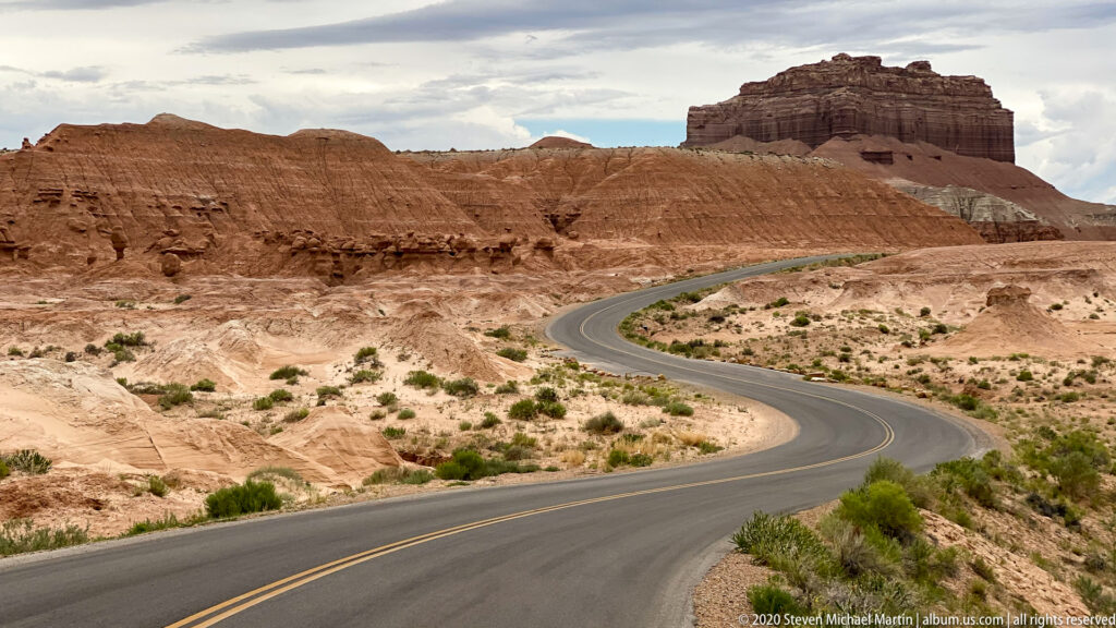A two-lane road curves through pale badlands beneath a blocky red mesa, with a car on the bend.