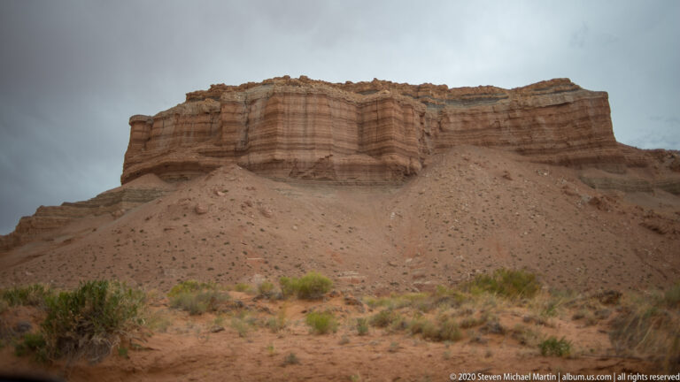 A broad layered sandstone cliff rises above a gravelly slope with scattered shrubs.