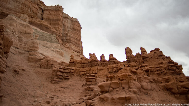 Narrow sandstone pinnacles form a jagged ridge beneath a tall cliff and cloudy sky.