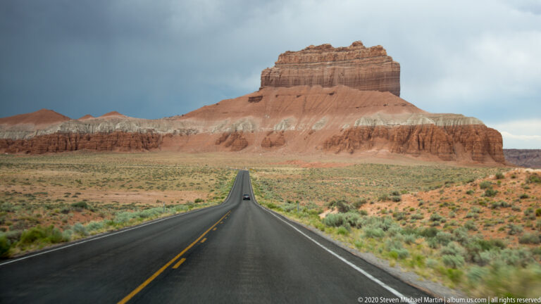 A straight road leads across desert scrub toward a large flat-topped sandstone butte.