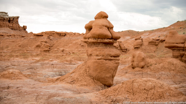 A tall lobed sandstone cap balances on a narrow, layered pedestal above shallow erosion channels.